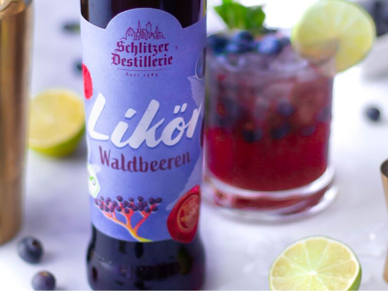Flasche Waldbeerenlikör und Cocktail mit Limette und Blaubeeren auf weißem Hintergrund.