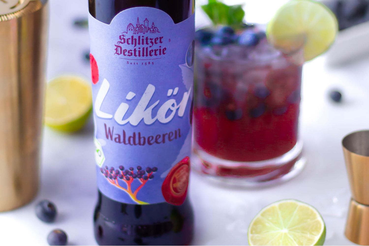 Flasche Waldbeerenlikör und Cocktail mit Limette und Blaubeeren auf weißem Hintergrund.