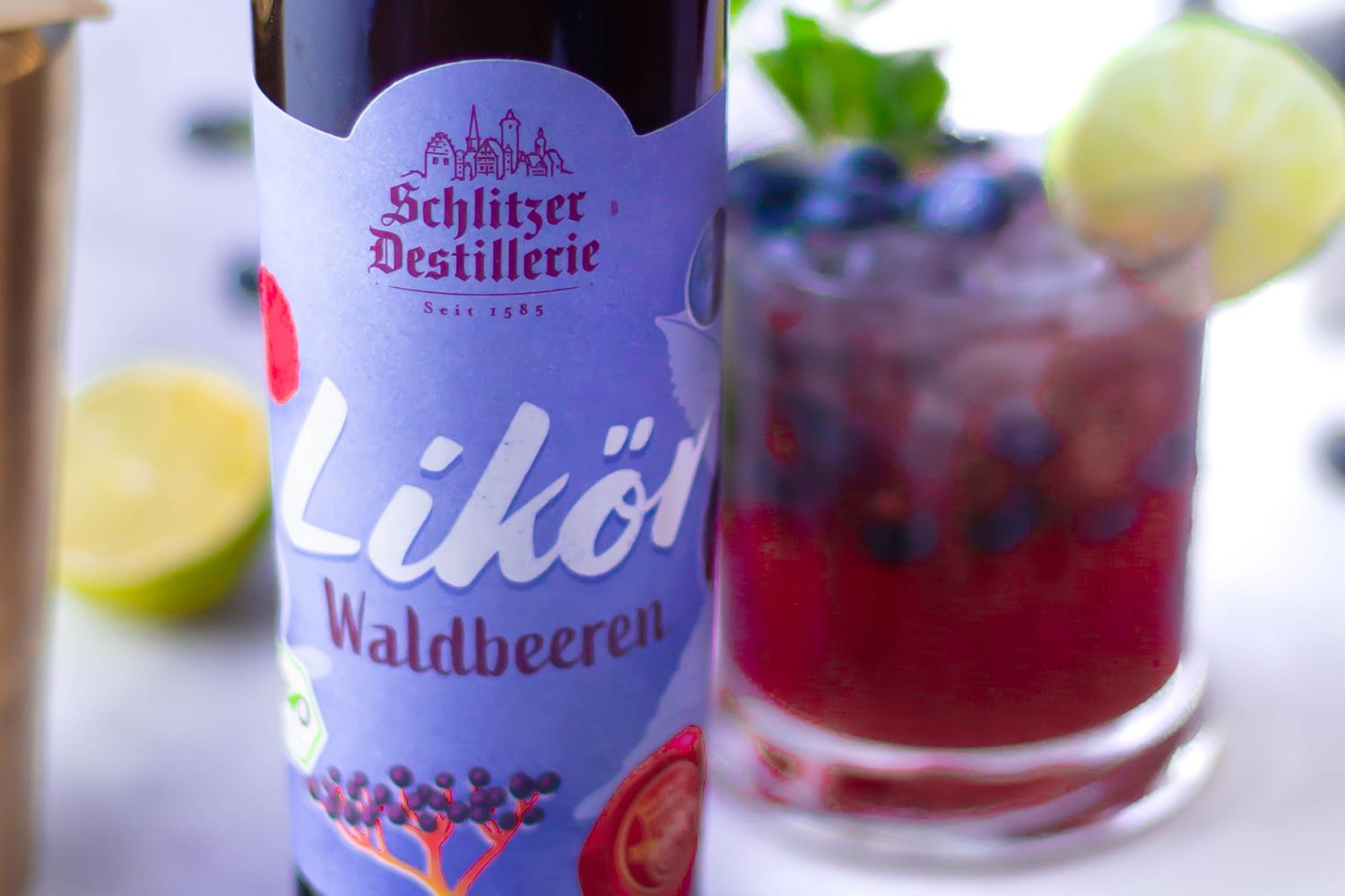 Blueberry Mule Flasche Likör mit Waldbeeren von Schlitzer Destillerie, ein Getränk mit Heidelbeeren und Limettenscheibe im Hintergrund.