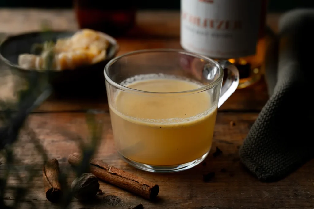 Tasse mit heißem Getränk auf Holztisch, umgeben von Zimtstangen und Gewürzen. Im Hintergrund unscharf Nüsse und Stoff.
