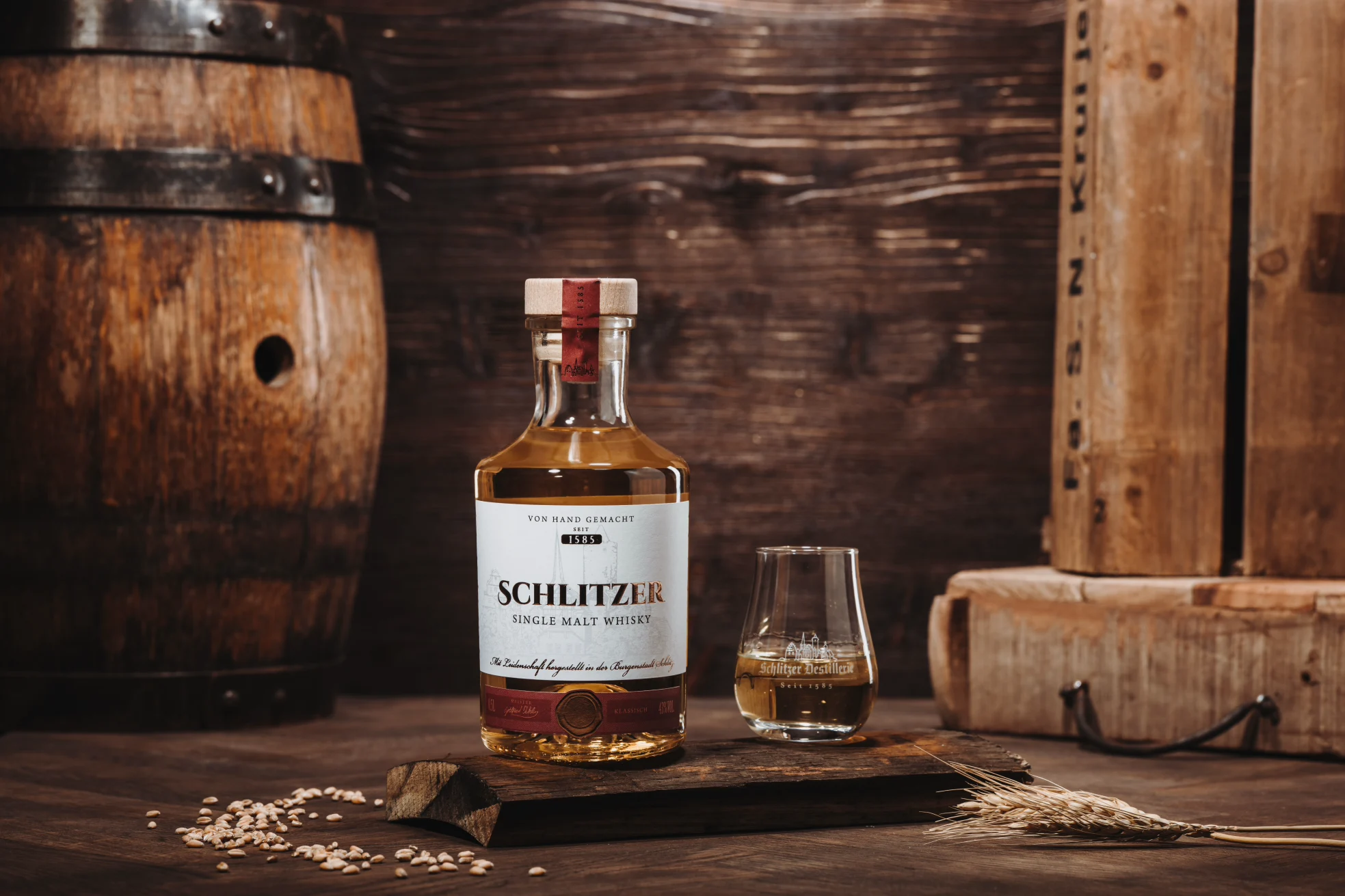 Whiskyflasche und Glas auf Holzuntergrund, daneben Getreide und Fass. Hintergrund rustikal mit Holzkisten.