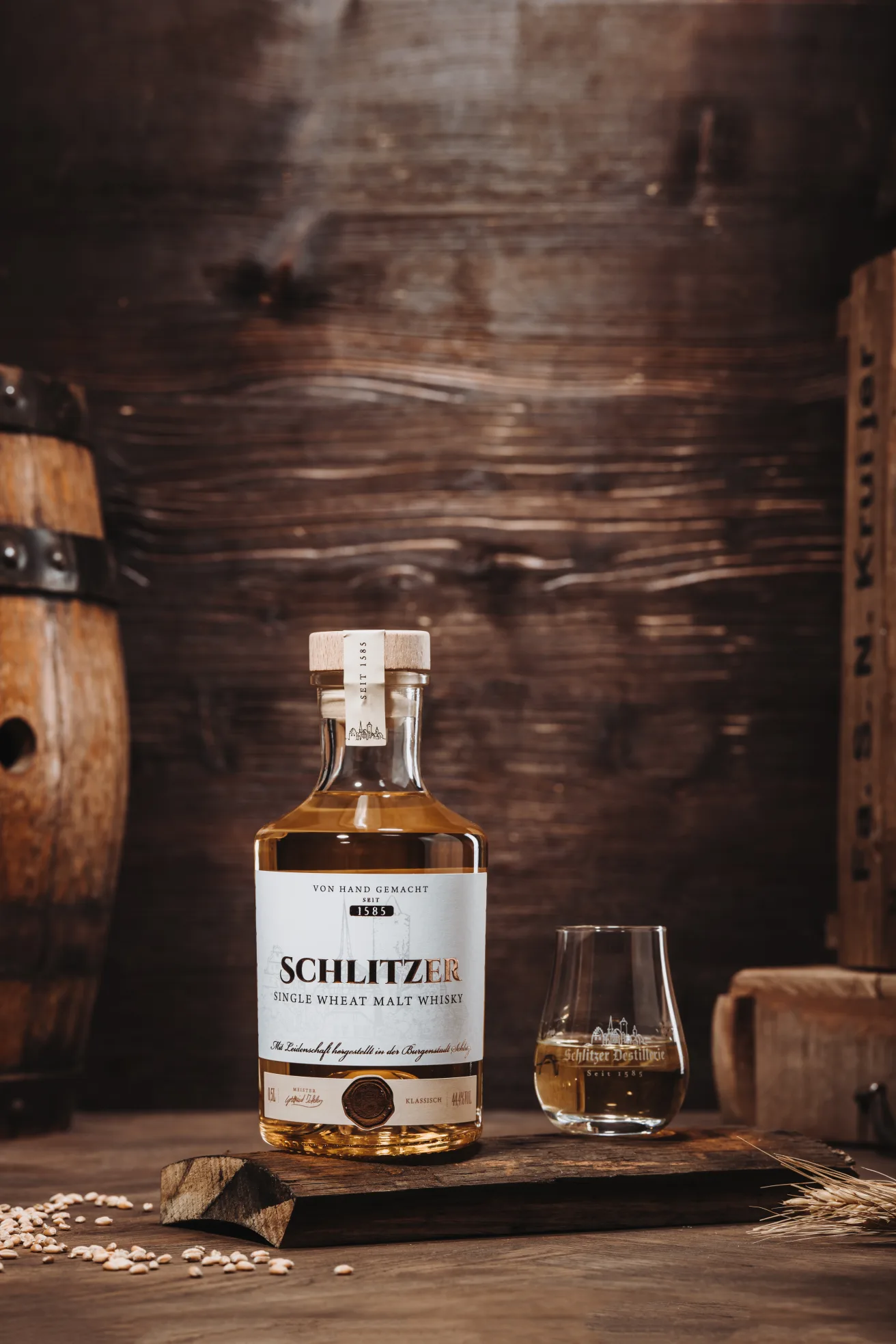 Flasche Schlitzer Whisky und Glas auf Holzbrett, umgeben von Gerstenkörnern, vor rustikalem Holzhintergrund.