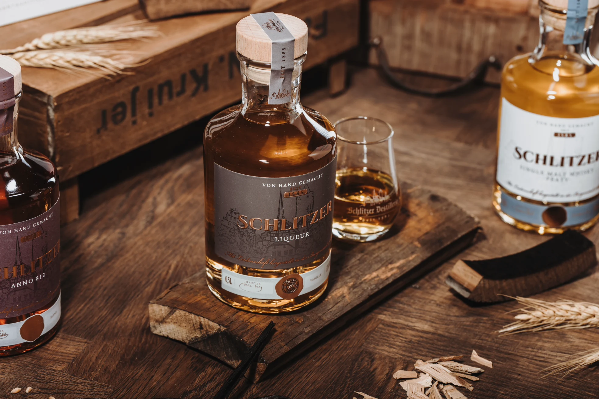 Flaschen und Glas mit „Schlitzer“ Likör auf Holzbrett, umgeben von Weizenähren und Holzstücken.