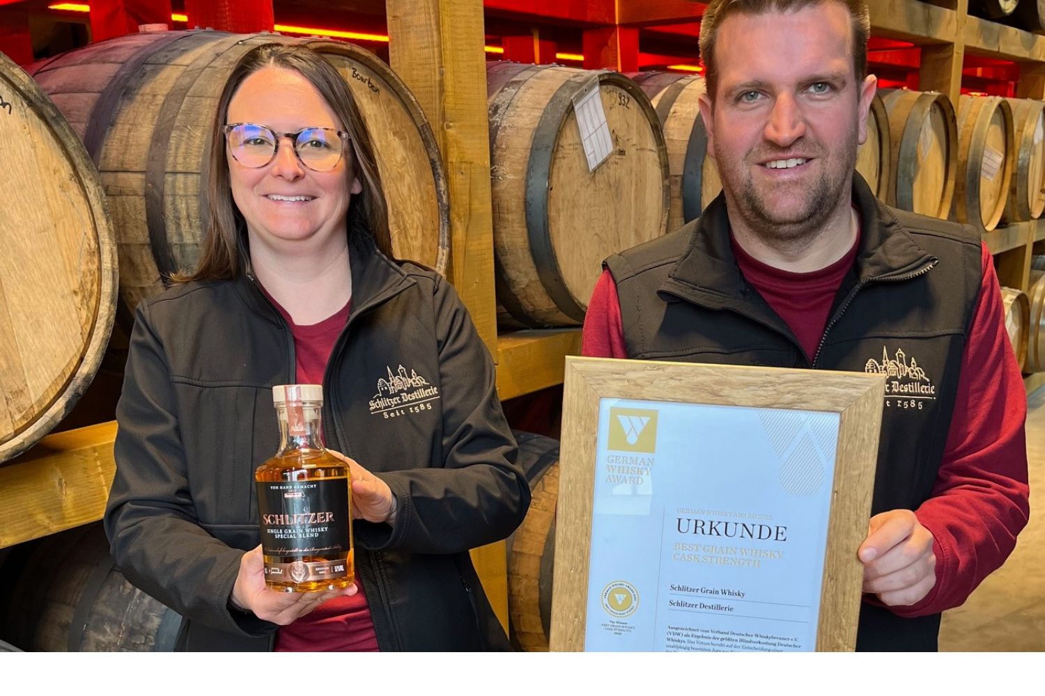 Zwei Personen präsentieren eine Whiskyflasche und eine Auszeichnungsurkunde vor Whiskyfässern in einer Destillerie.