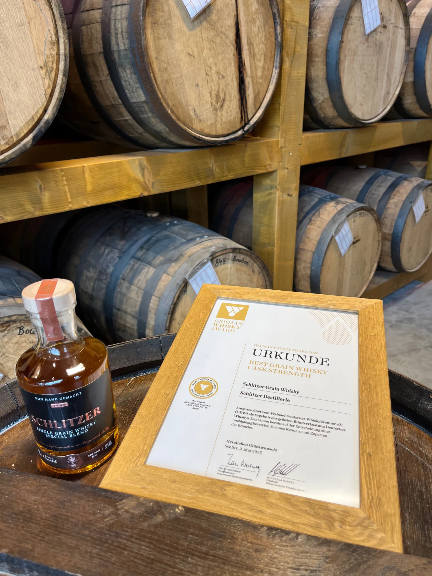 Grain Whisky Cask Strength Whiskyflasche und Auszeichnungsurkunde auf einem Fass vor einem Regal mit Holzfässern.