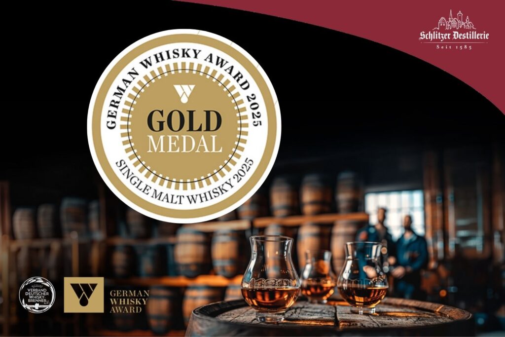 Goldmedaille für Single Malt Whisky 2025 vor Fasslager, Gläsern und zwei unscharfen Personen im Hintergrund.