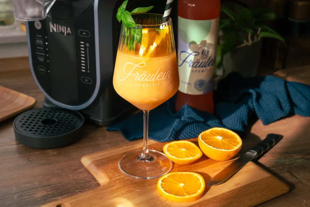 Ein Glas mit orangefarbenem Aperitif und Basilikum, daneben Orangen und ein Messer auf einem Schneidebrett.