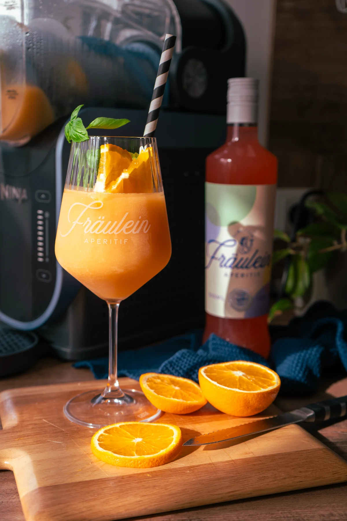 Fräulein Slushi Oranger Aperitif im Glas mit Basilikumblatt, daneben geschnittene Orangen auf Holzbrett, Flasche im Hintergrund.