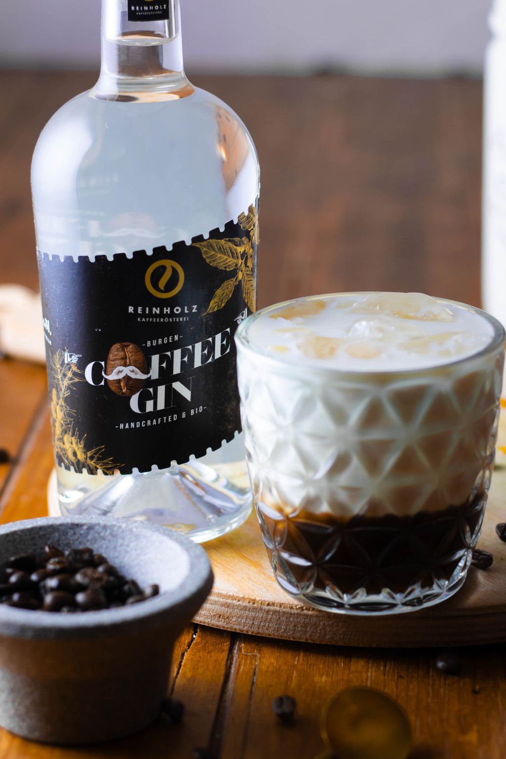 Gin White Russian Kaffeegin-Flasche neben Cocktail im Glas mit Eis und Sahne, auf Holzbrett serviert. Daneben Schale mit Kaffeebohnen.