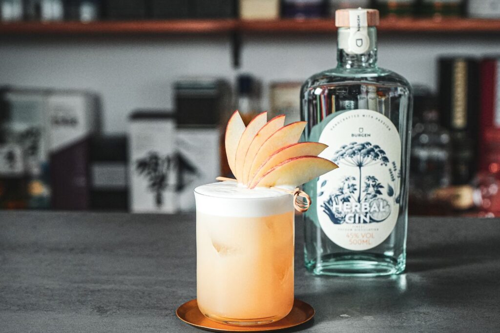 Ein Cocktail im Glas mit aufgeschäumtem Topping und Apfelscheiben-Garnitur vor einer Flasche Gin auf einer Theke.
