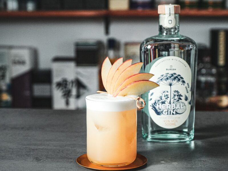 Ein Cocktail im Glas mit aufgeschäumtem Topping und Apfelscheiben-Garnitur vor einer Flasche Gin auf einer Theke.