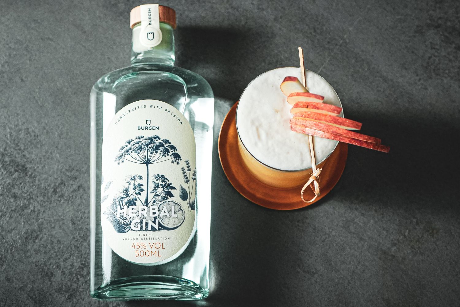 Ein Cocktail im Glas mit aufgeschäumtem Topping und Apfelscheiben-Garnitur vor einer Flasche Gin auf einer Theke.