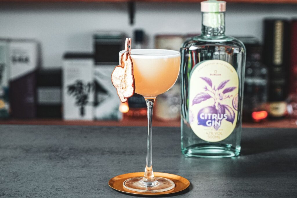 Cocktail in einem Glas mit getrockneter Birne am Rand, daneben eine Flasche Citrus Gin auf grauer Theke.