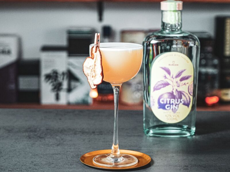 Cocktail in einem Glas mit getrockneter Birne am Rand, daneben eine Flasche Citrus Gin auf grauer Theke.