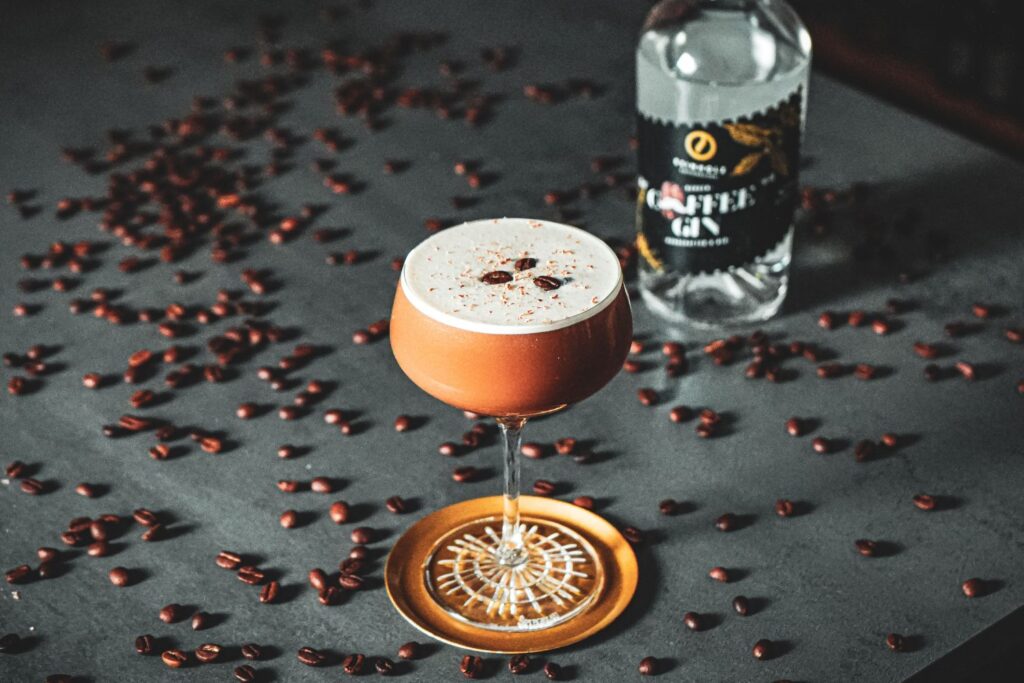 Cocktailglas mit Kaffee-Getränk und Schaum, dekoriert mit Kaffeebohnen. Im Hintergrund Kaffeelikörflasche und Bohnen.