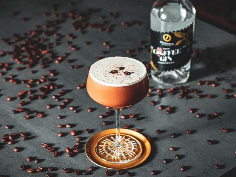 Cocktailglas mit Kaffee-Getränk und Schaum, dekoriert mit Kaffeebohnen. Im Hintergrund Kaffeelikörflasche und Bohnen.