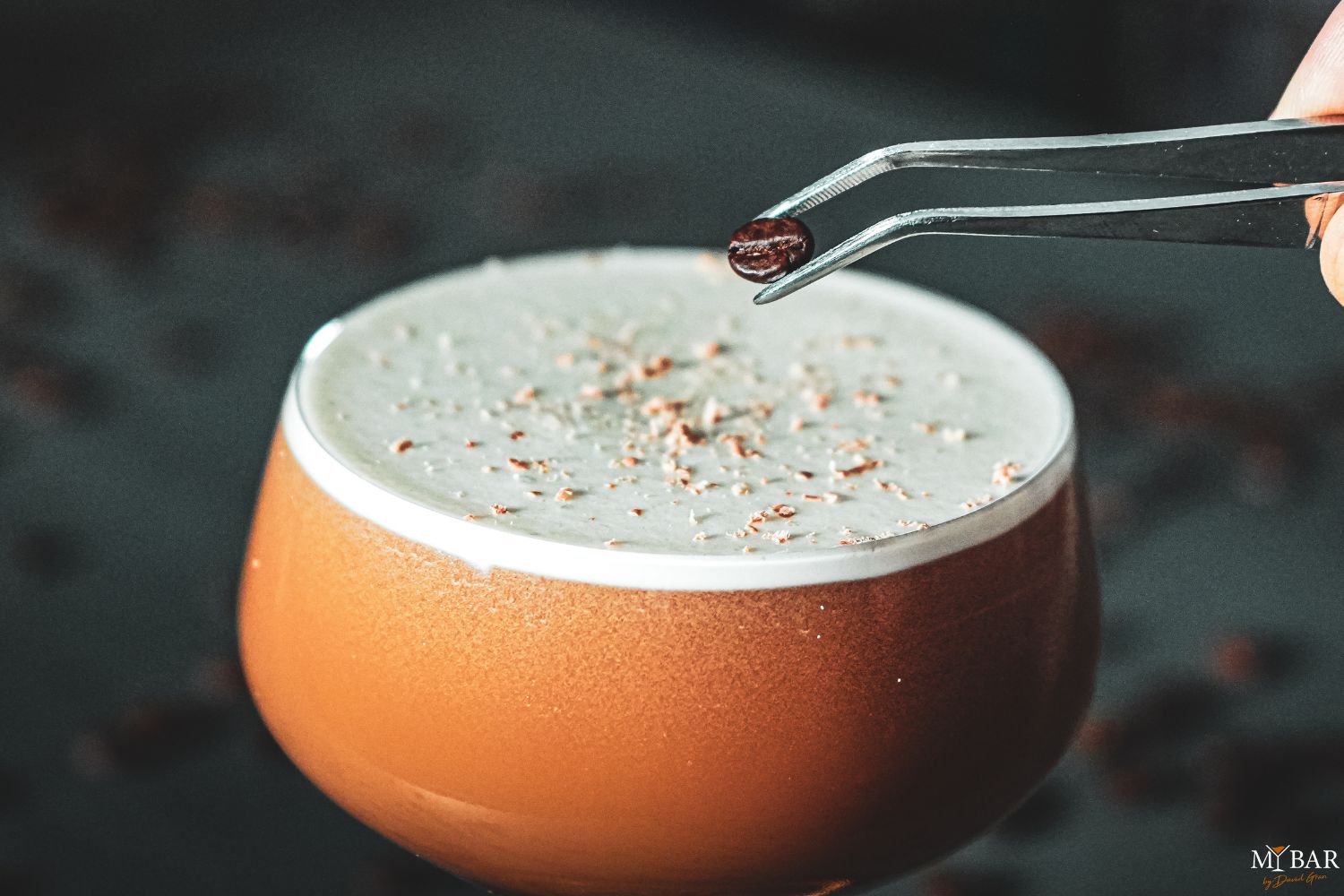 Nahaufnahme eines Getränks mit Schaum, das mit einer Pinzette mit einer Kaffeebohne garniert wird.