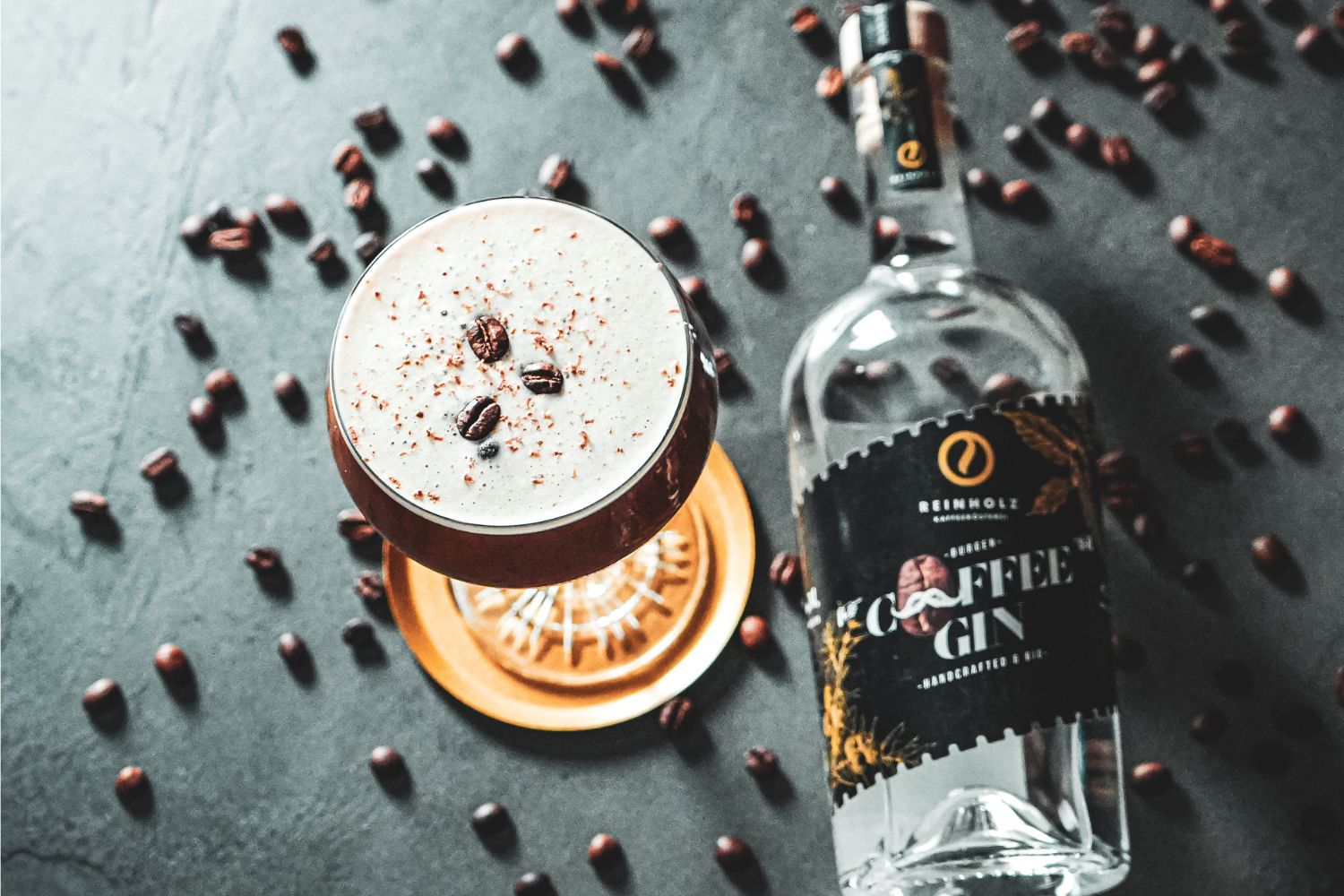 Cocktailglas mit Kaffeedrink, garniert mit Bohnen, neben Coffee-Gin-Flasche auf grauer Oberfläche, umgeben von Kaffeebohnen.