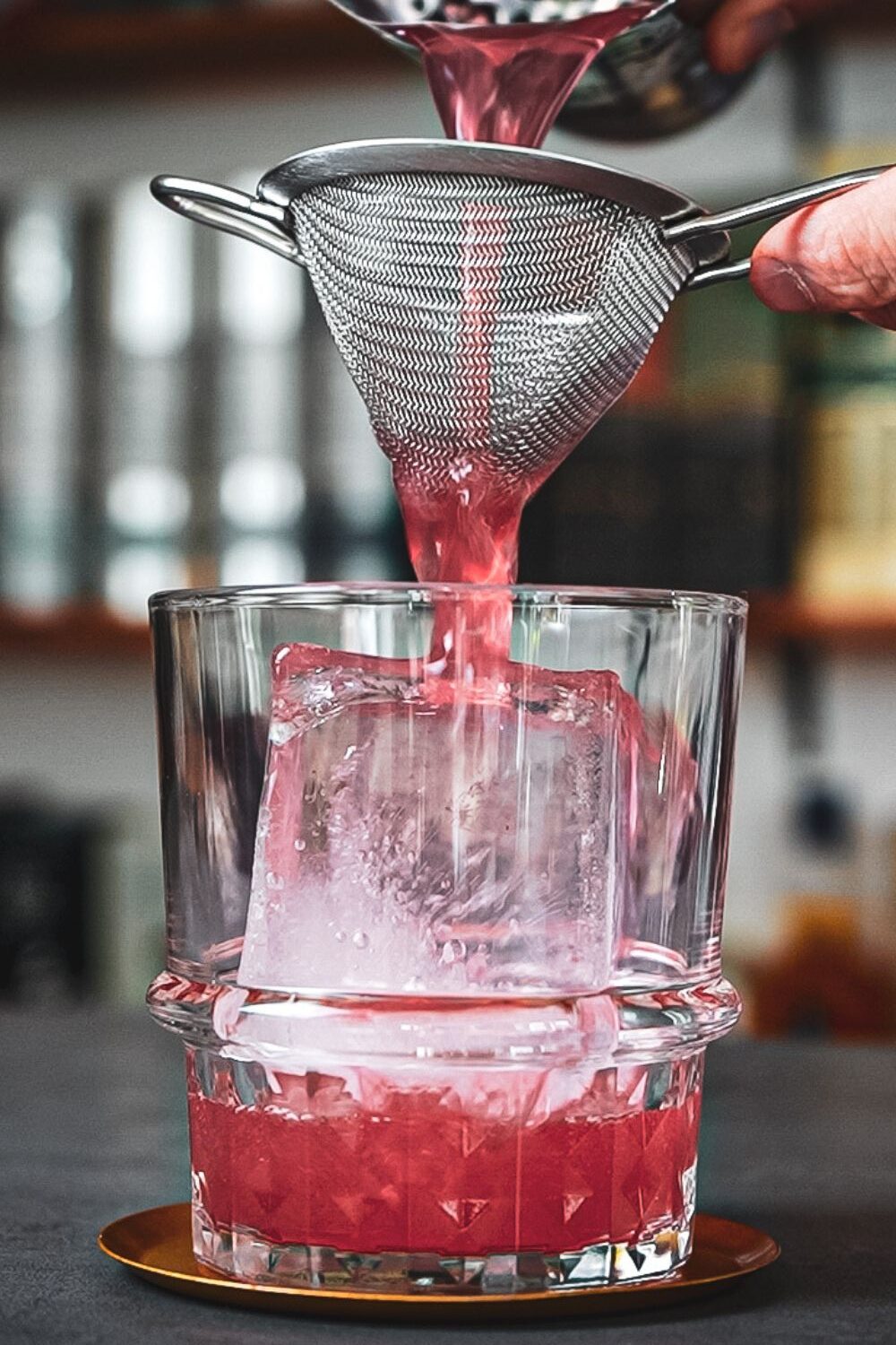Iced Tea on the Castle Rosa Cocktail wird durch ein Sieb in ein Glas mit großem Eiswürfel gegossen.