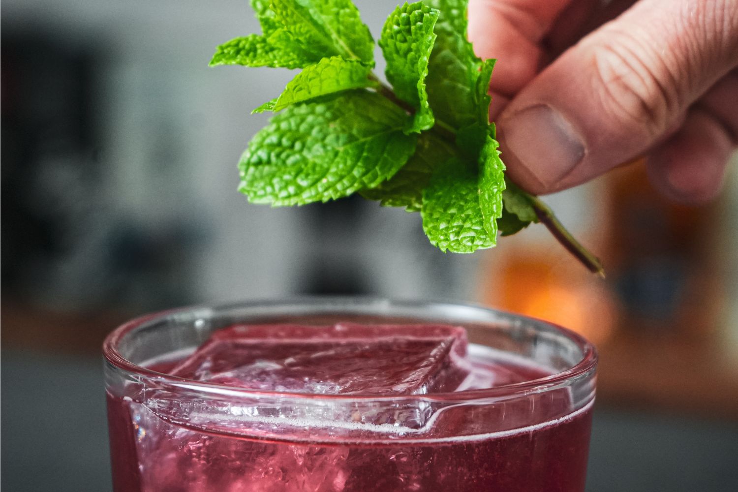 Iced Tea on the Castle Hand hält frische Minze über einem Glas mit violettem Getränk und Eiswürfeln.