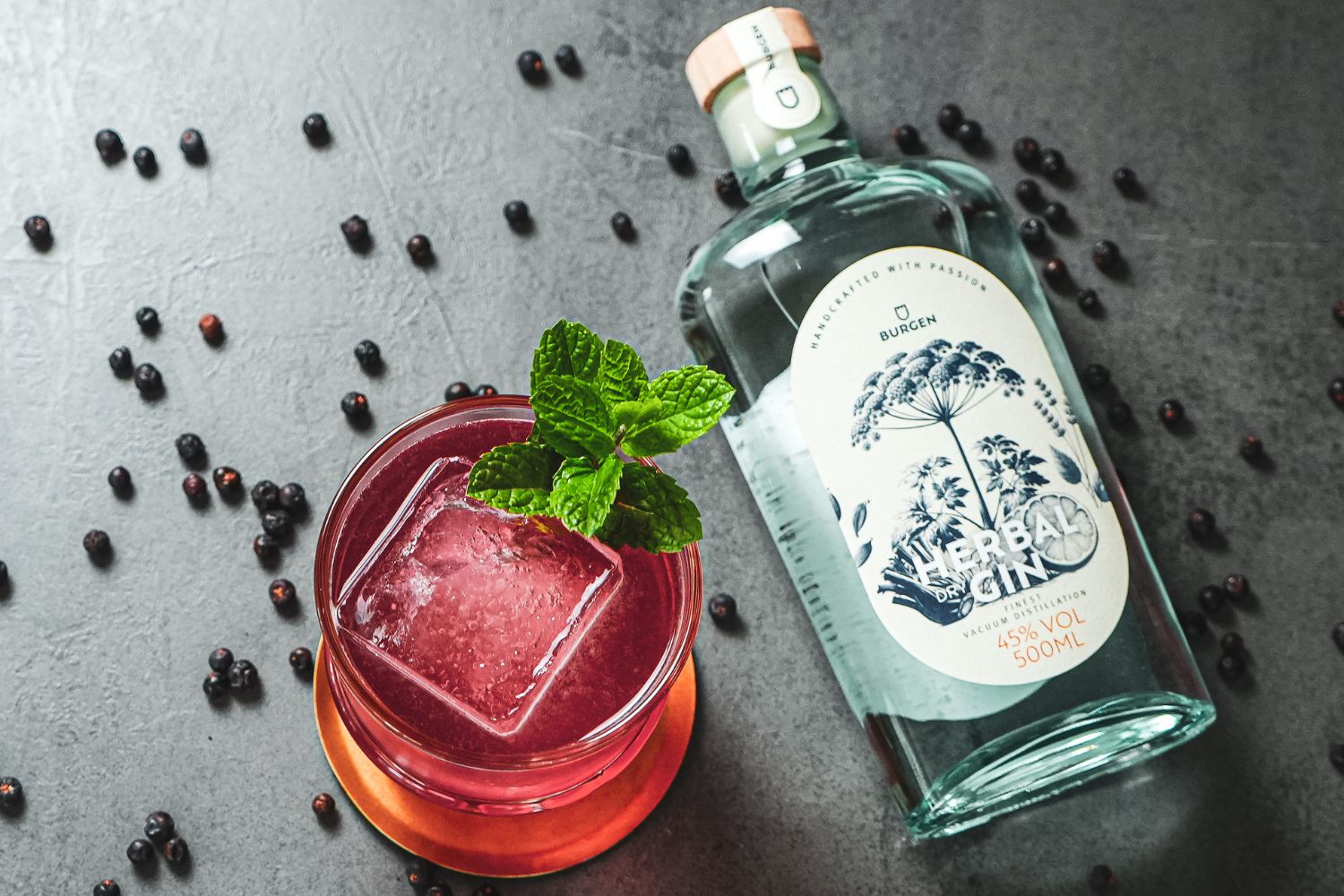 Iced Tea on the Castle Rotes Getränk mit Eis und Minze neben einer Flasche Gin auf grauem Untergrund, umgeben von Wacholderbeeren.