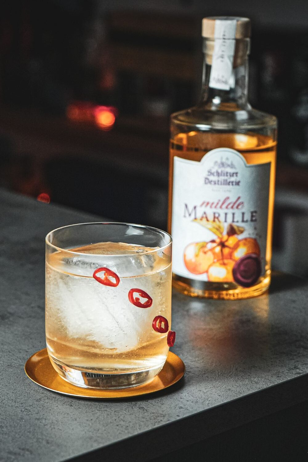 Cocktailglas mit Eis und Chilischoten sitzt auf einem goldenen Untersetzer neben einer Flasche Marillenlikör.