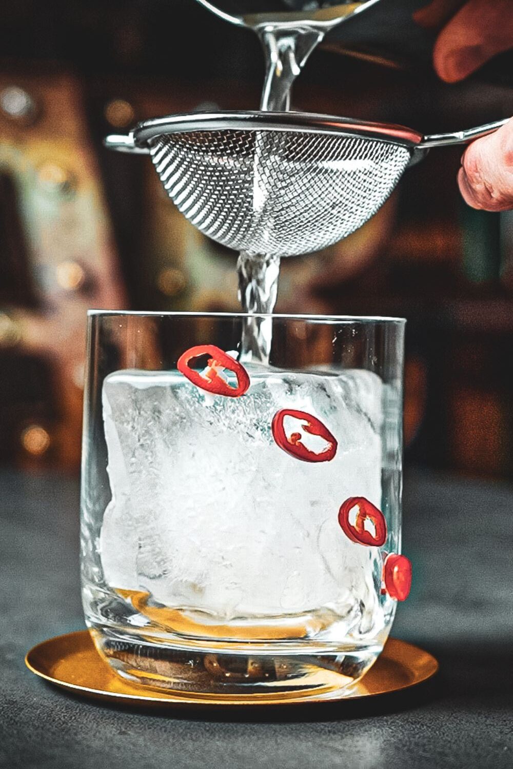 Ein Getränk wird durch ein Sieb in ein Glas mit Eiswürfel und Chilischeiben gegossen, das auf einem goldenen Untersetzer steht.