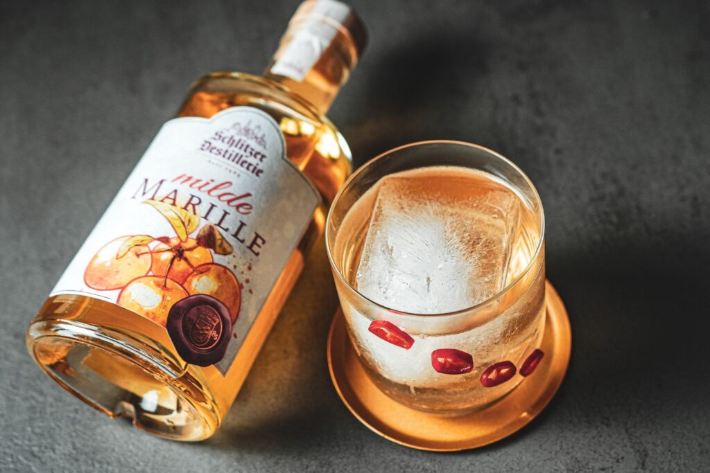Flasche Marillenschnaps und Glas mit Eiswürfel und rotem Dekor auf Untersetzer.