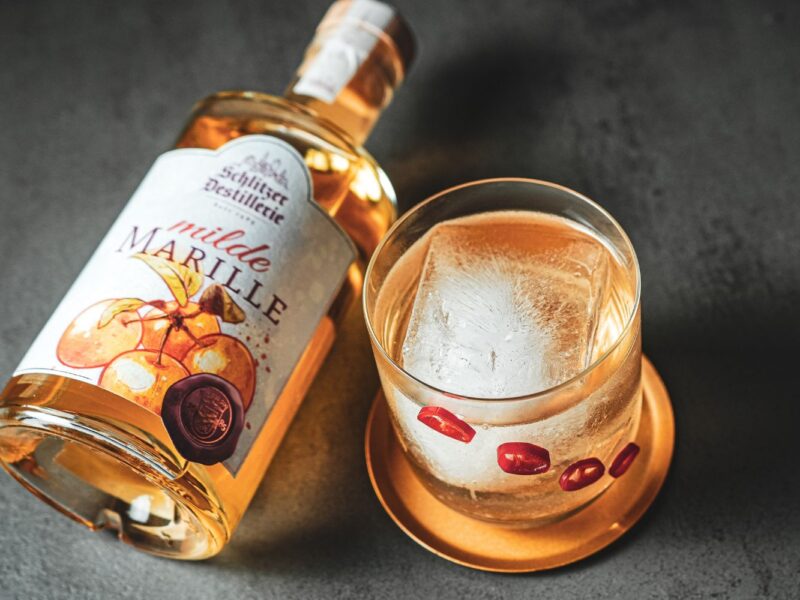 Flasche Marillenschnaps und Glas mit Eiswürfel und rotem Dekor auf Untersetzer.