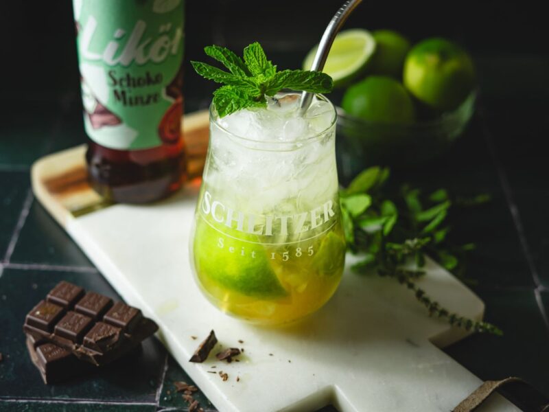 Cocktailglas mit Minze, Limetten und Eis auf Schneidebrett. Neben Schoko, Likörflasche und Limette im Hintergrund.