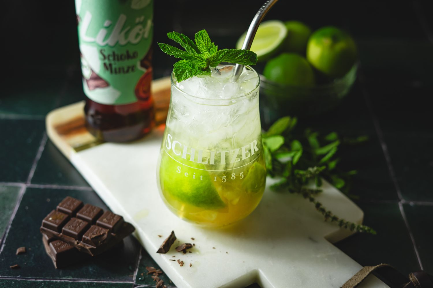 Chocolate Mint Mojito Cocktailglas mit Minze, Limetten und Eis auf Schneidebrett. Neben Schoko, Likörflasche und Limette im Hintergrund.