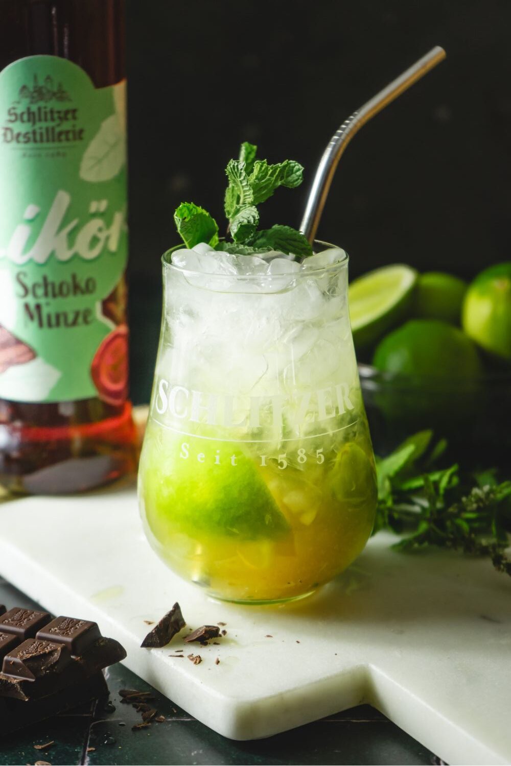 Glas mit Minz-Cocktail, Eis, Limette und Metallstrohhalm, daneben Schoko-Minze-Likörflasche und Schokolade.