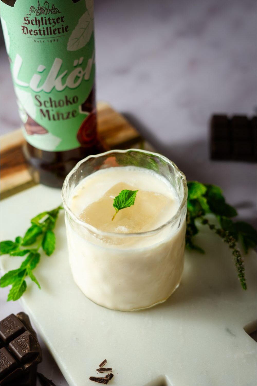 Glas mit cremigem Schoko-Minze-Drink, garniert mit Minzblatt, Flasche im Hintergrund, Schokostückchen daneben.