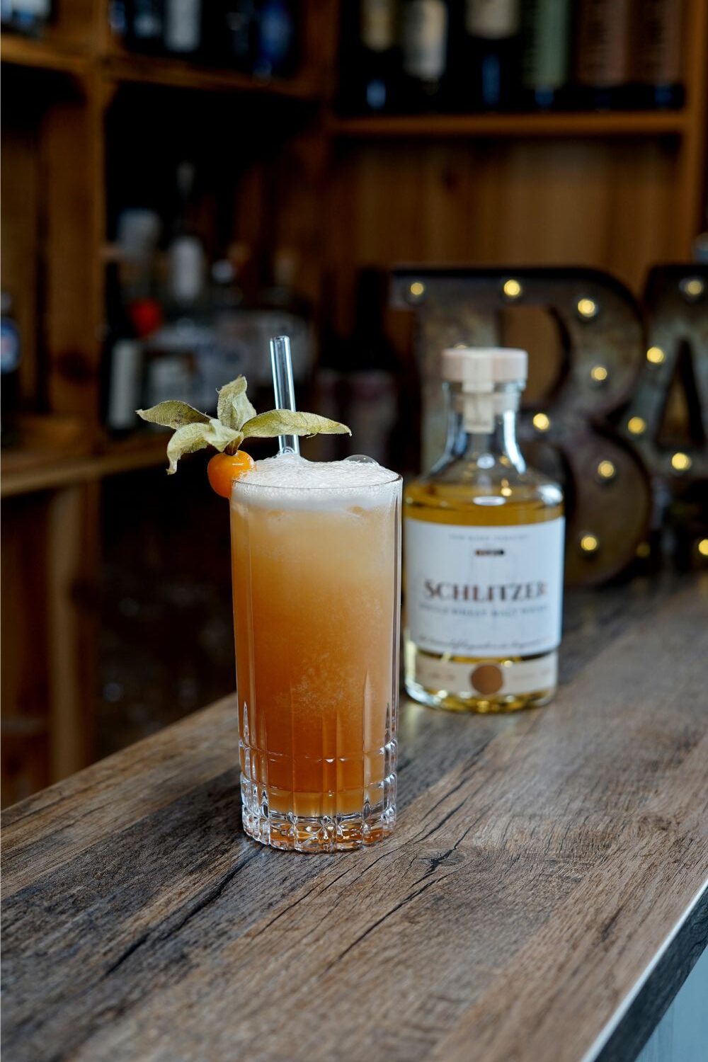 Eye of the Storm Hoher Cocktail mit orangefarbenem Getränk und Physalis-Garnitur auf Holztresen, im Hintergrund eine Flasche Schlitzer.
