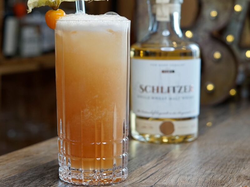 Ein cremiger Cocktail mit Physalis-Deko steht vor einer Whiskyflasche auf einem Holztisch.