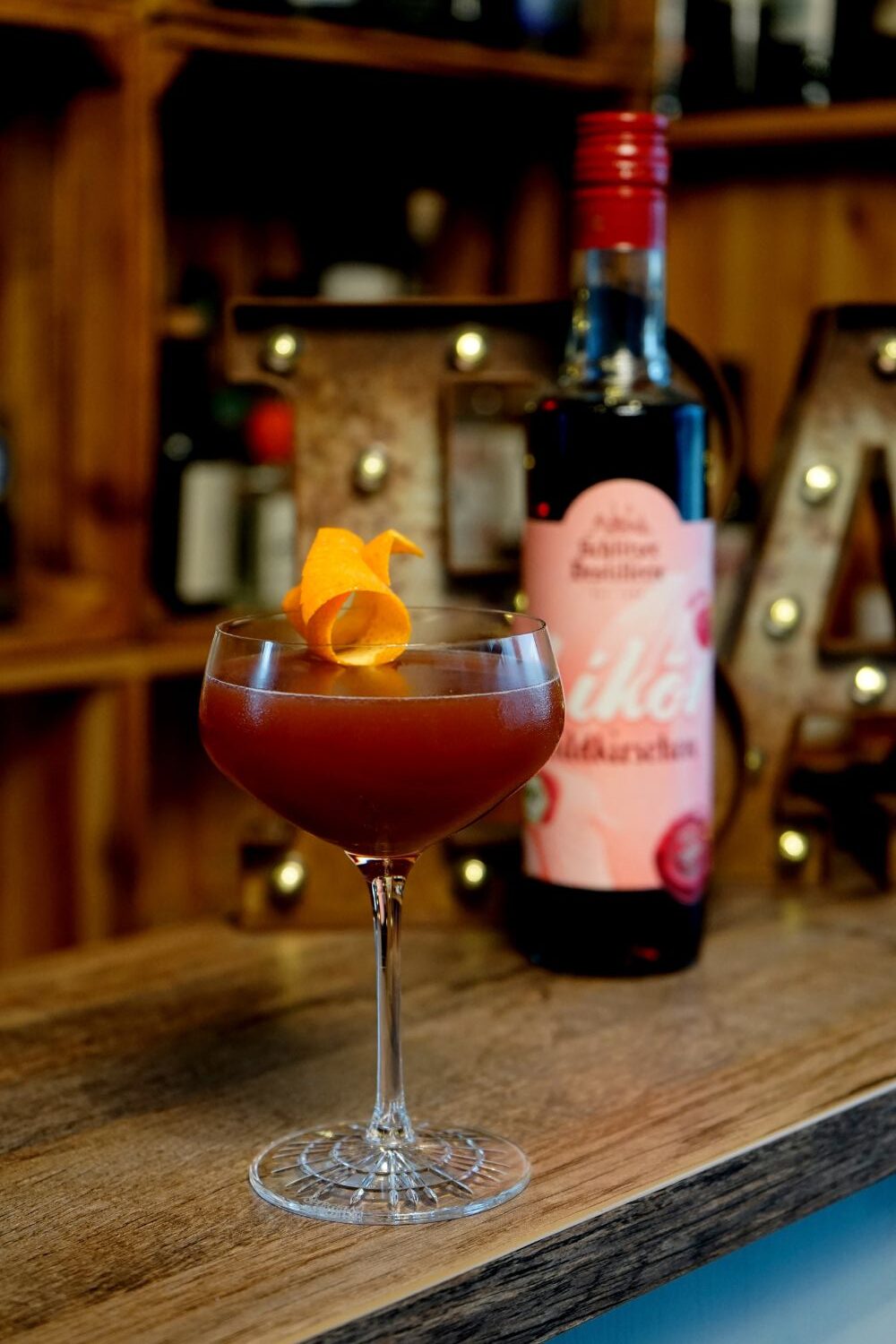 Schlitzer Blood and Sand Cocktail mit Orangenschale garniert, steht auf Theke. Dahinter Flasche mit rotem Etikett. Hintergrund unscharf.