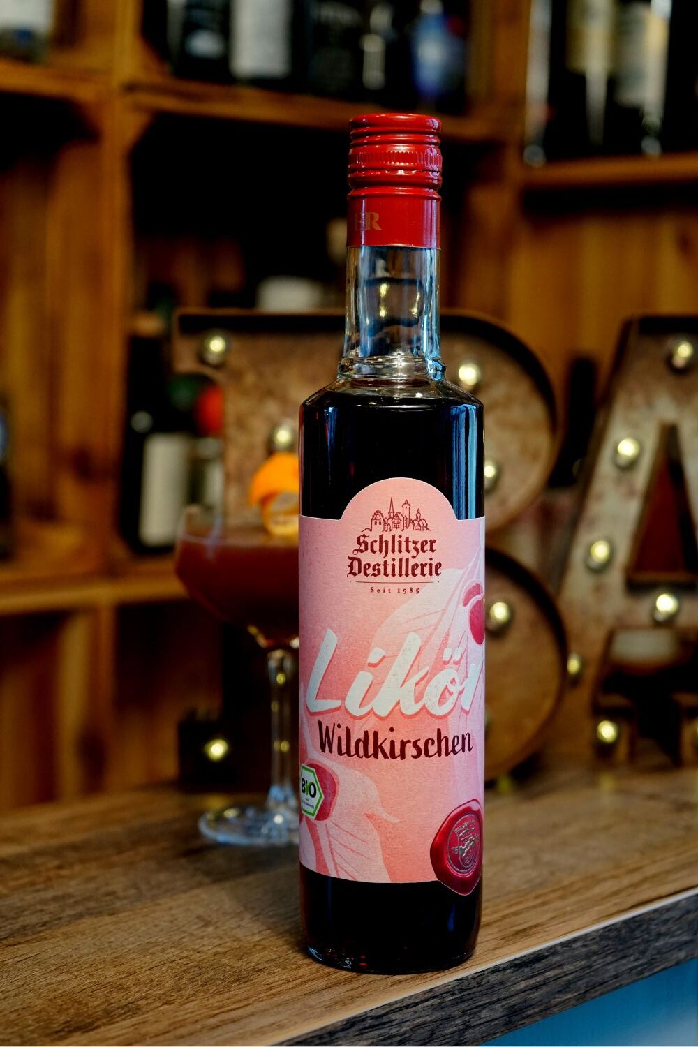 Schlitzer Blood and Sand Flasche Wildkirschlikör von Schlitzer Destillerie auf einem Holztresen, im Hintergrund ein Glas und Regale.