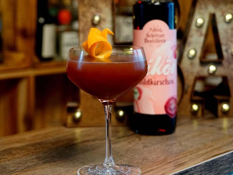 Cocktail im Glas mit einer Orangenschale garniert, dahinter eine Flasche Likör auf einer Holztheke.