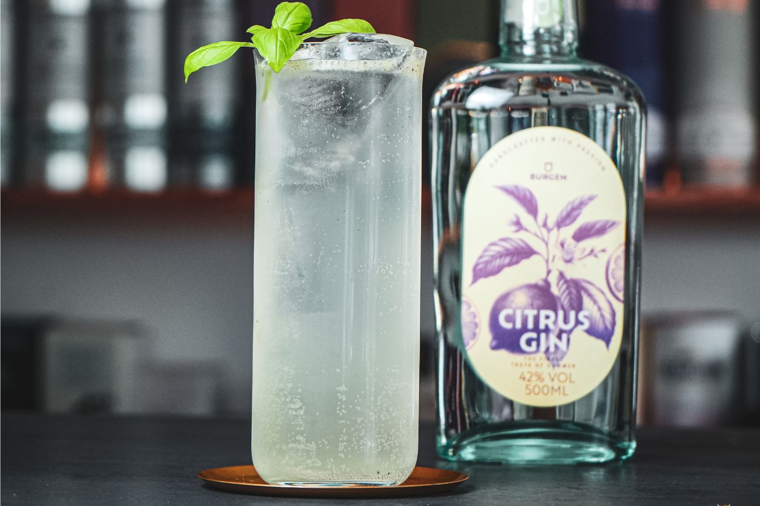 Hohes Glas mit klarem, sprudelndem Getränk und Basilikum, daneben eine Flasche Citrus Gin auf einem Tisch.