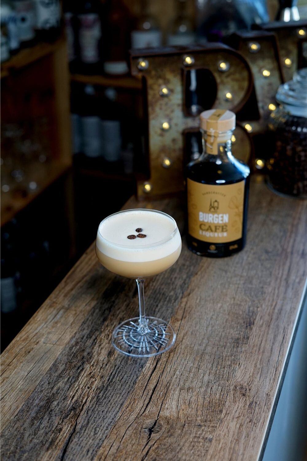 Toasted Almond Cocktail im Kelchglas auf Holztresen, garniert mit Kaffeebohnen. Im Hintergrund eine Flasche Burgen Kaffee Likör.