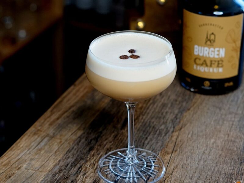 Kaffeecocktail in einem Glas mit drei Kaffeebohnen, neben einer Flasche Burgen Café Likör auf einem Holztisch.