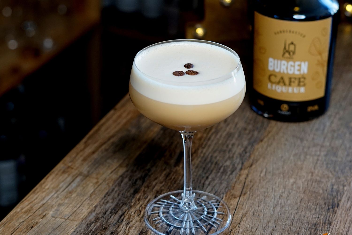 Kaffeecocktail in einem Glas mit drei Kaffeebohnen, neben einer Flasche Burgen Café Likör auf einem Holztisch.