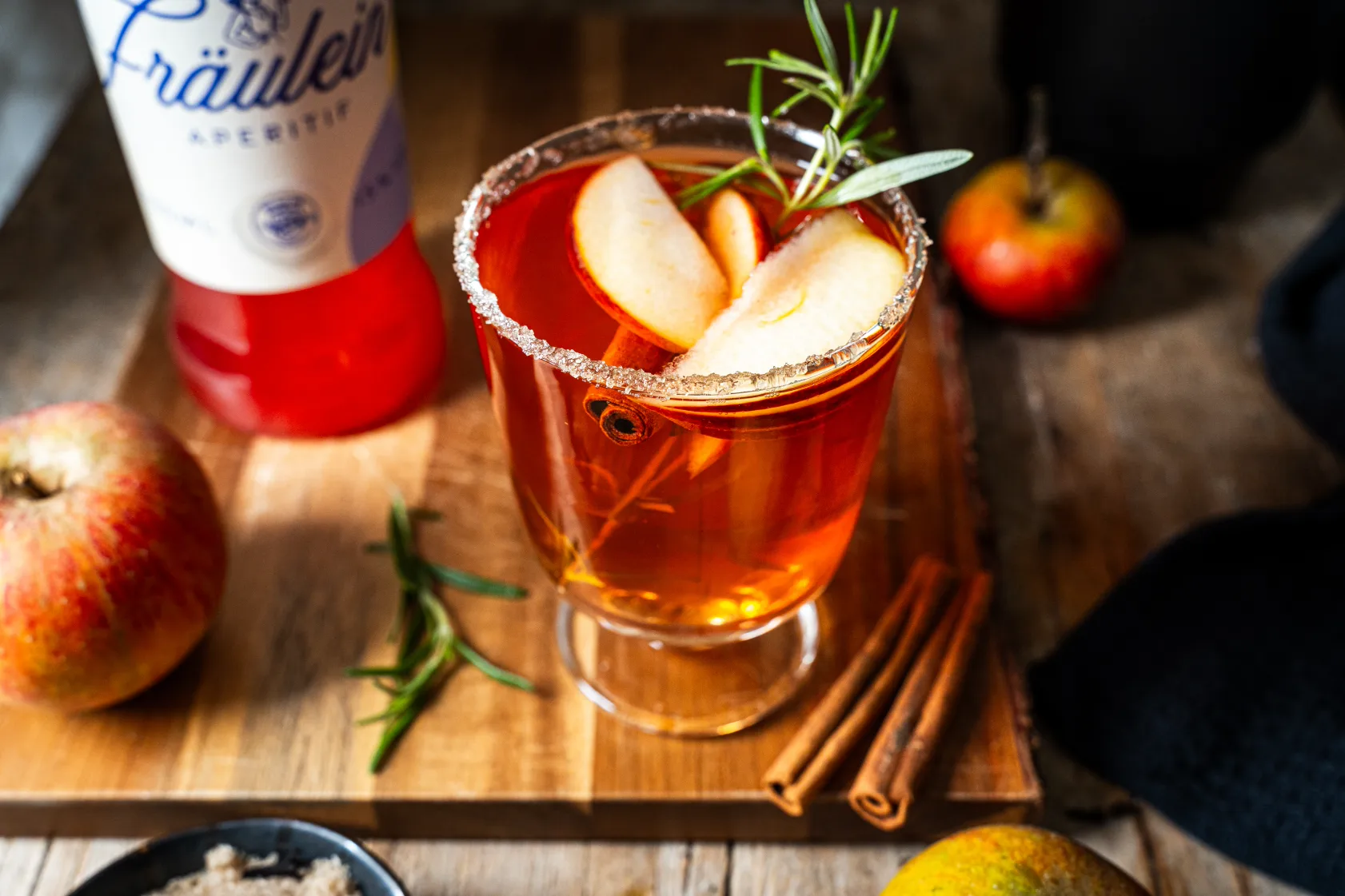 Ein Glas Cocktail mit Apfelscheiben, Rosmarin und Zimtstange. Im Hintergrund Äpfel und eine rote Flasche.