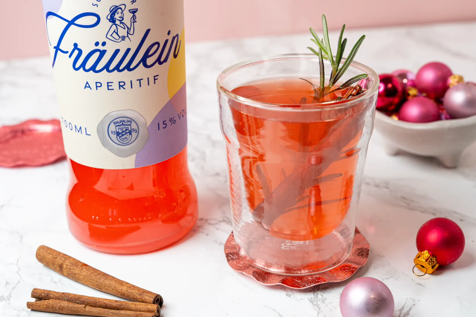 Schlitzer_Fraeulein_Hot_Punch (3 von 6)1 Flasche Fräulein Aperitif und Glas mit rosa Getränk auf Mamortisch, Zimtstangen, Rosmarin und rote Kugeln als Dekoration.