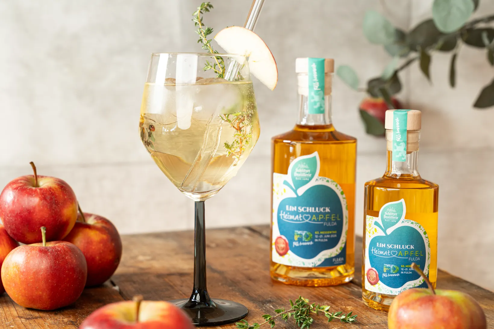 Weinglas mit Apfelgetränk, Apfelscheibe, Thymian und zwei Apfellikörflaschen. Rote Äpfel als Dekoration.