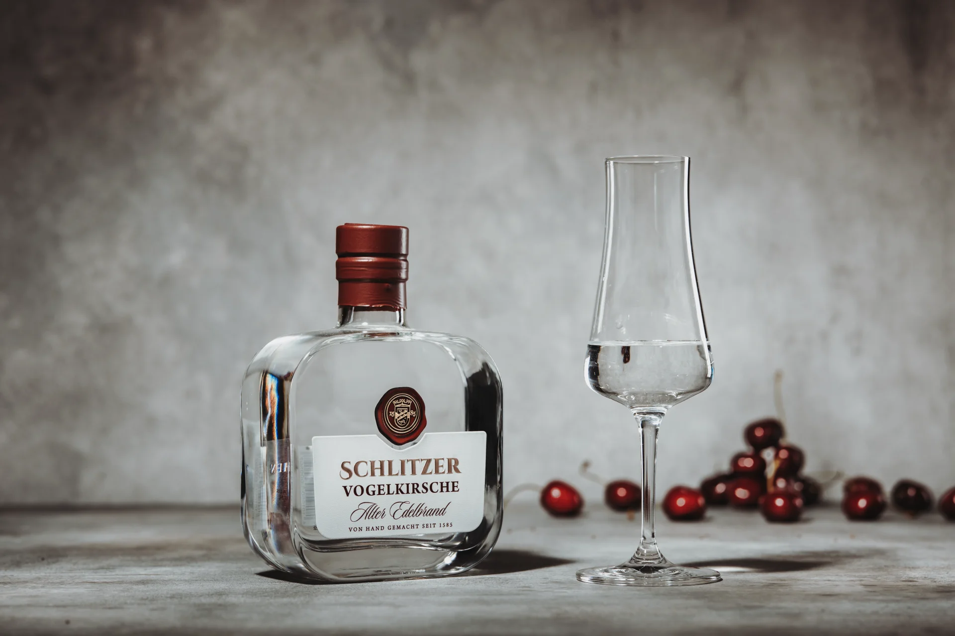 Vogelkirschenbrand in eleganter 0,5L-Flasche – Edler Obstbrand aus der Schlitzer Destillerie Klare Glasflasche mit Vogelkirschen-Brand der Schlitzer Destillerie, Etikett mit rotem Logo und goldener Schrift, daneben frische Vogelkirschen und Holzuntergrund – hochwertiger deutscher Obstbrand.