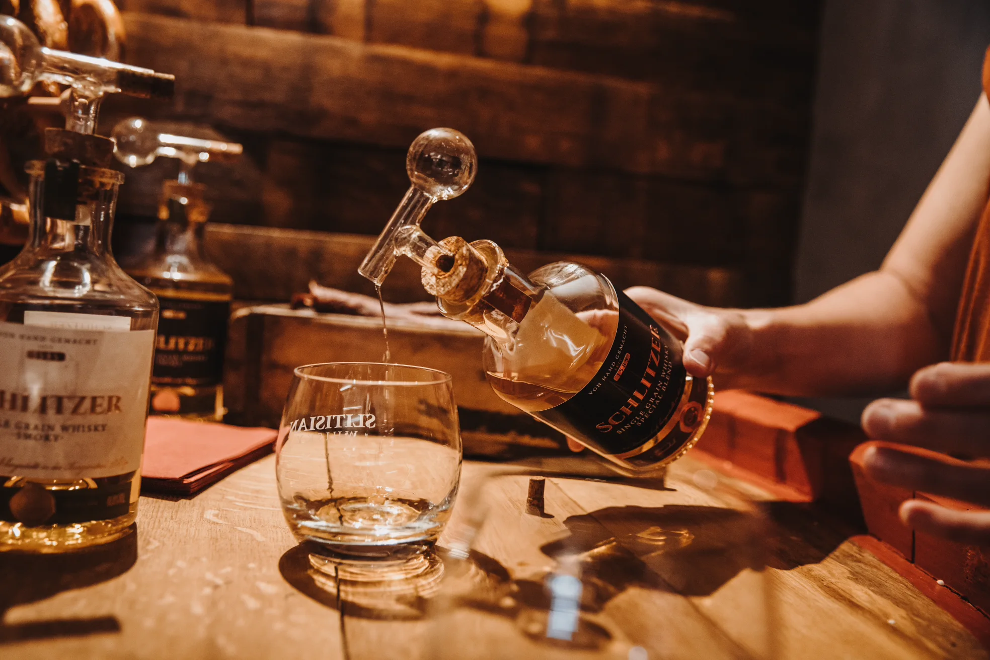 Whisky-Tasting in der Schlitzer Destillerie â Genuss in historischer AtmosphĂ€re Drei Personen sitzen in einer rustikalen Brennerei an einem Holztisch und verkosten verschiedene Spirituosenproben unter warmem Licht, im Hintergrund sind Kupferbrennblasen und HolzfĂ€sser sichtbar â Tasting in der Schlitzer Destillerie.