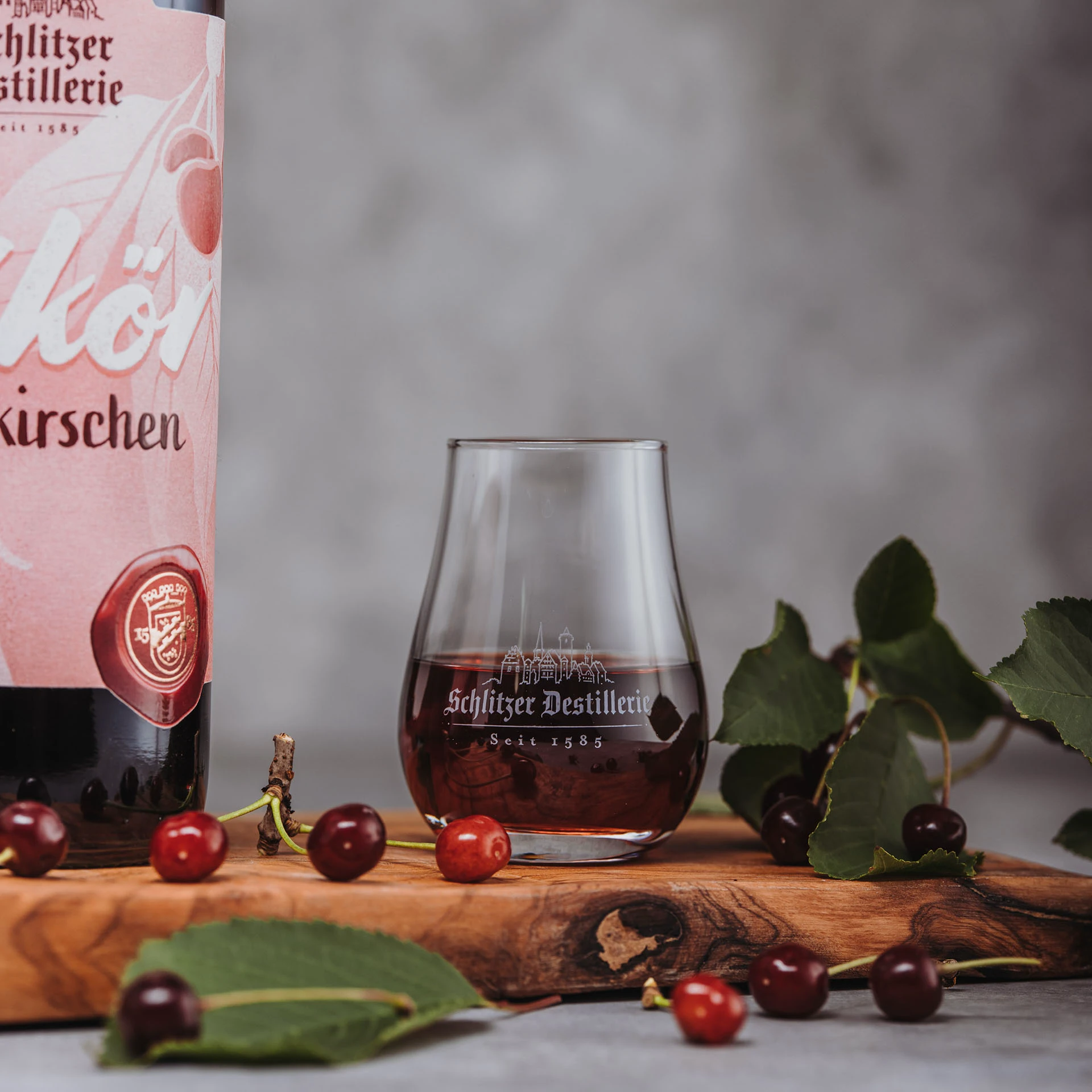 Schlitzer Wilkirschen Likör – Edler Kirschlikör in hochwertiger 0,5L Flasche Flasche Schlitzer Wilkirschen Likör mit rotem Etikett und goldener Schrift auf weißem Hintergrund, daneben zwei reife Wildkirschen – hochwertiger Wildkirsch-Likör aus der Schlitzer Destillerie.