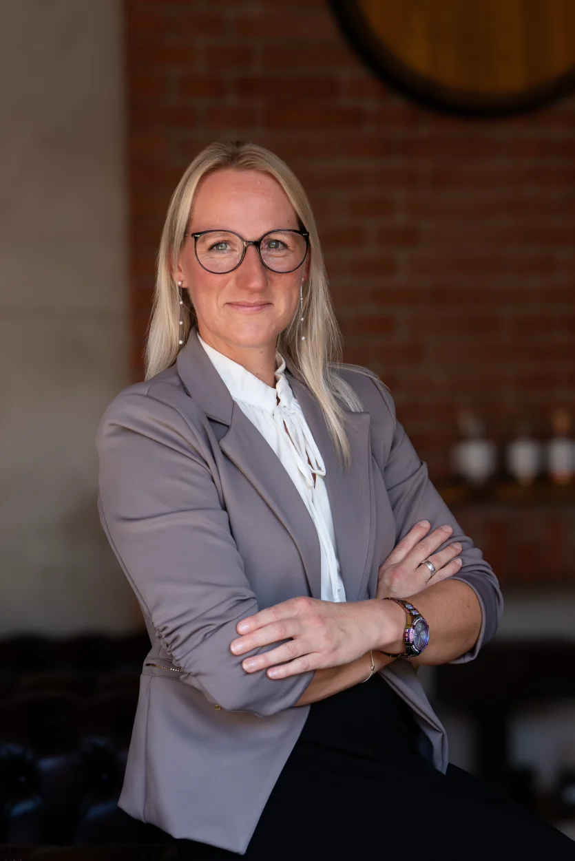 Vertriebsmitarbeiterin Jennifer Hartung der Schlitzer Destillerie im Portrait Jennifer Hartung, Vertriebsmitarbeiterin der Schlitzer Destillerie, lächelt in businessgerechter Kleidung vor einem Hintergrund mit edlen Spirituosen – professionelle Porträtaufnahme für Unternehmenspräsentation.