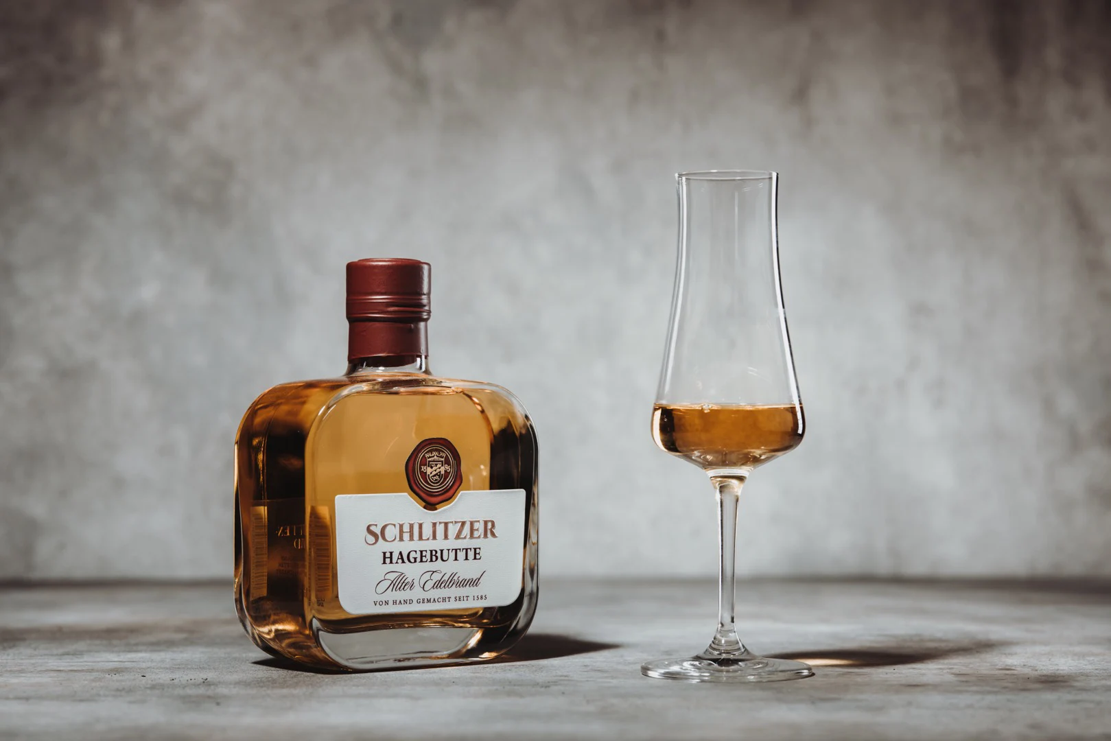 Hagebutte Edelbrand – Hochwertiger Fruchtbrand aus reifen Wildfrüchten Flasche Schlitzer Hagebutte Edelbrand mit elegantem Etikett auf hellem Untergrund, daneben frische Hagebutten – hochwertiger Fruchtbrand aus Deutschland.